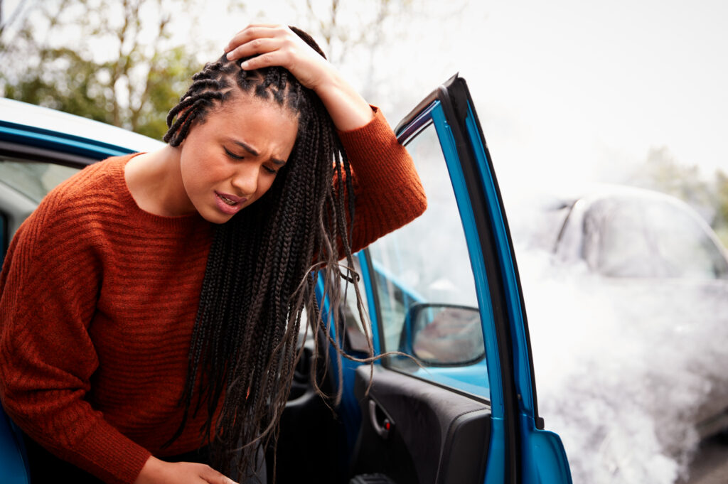 A woman exits her car after a car accident, holding her head in pain. 