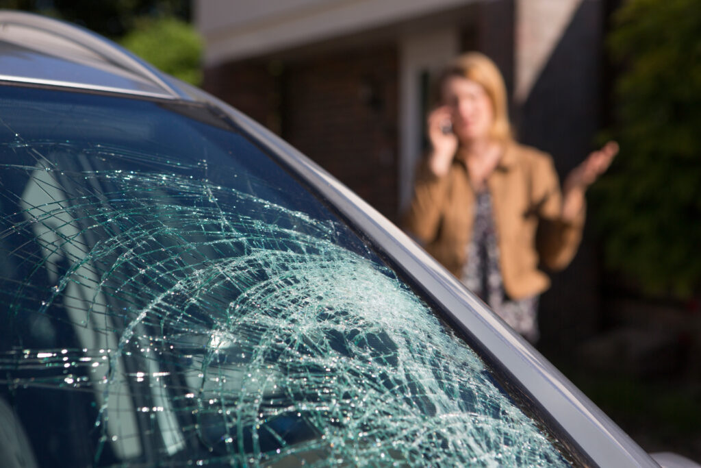 A woman stands outside of her car with a broken windshield after an auto accident. She is making a telephone call.