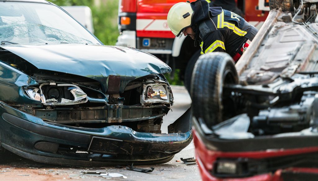 A first responder assesses the damage of a car crash between two vehicles. One has been flipped over and the other car has heavy damage to the front end.