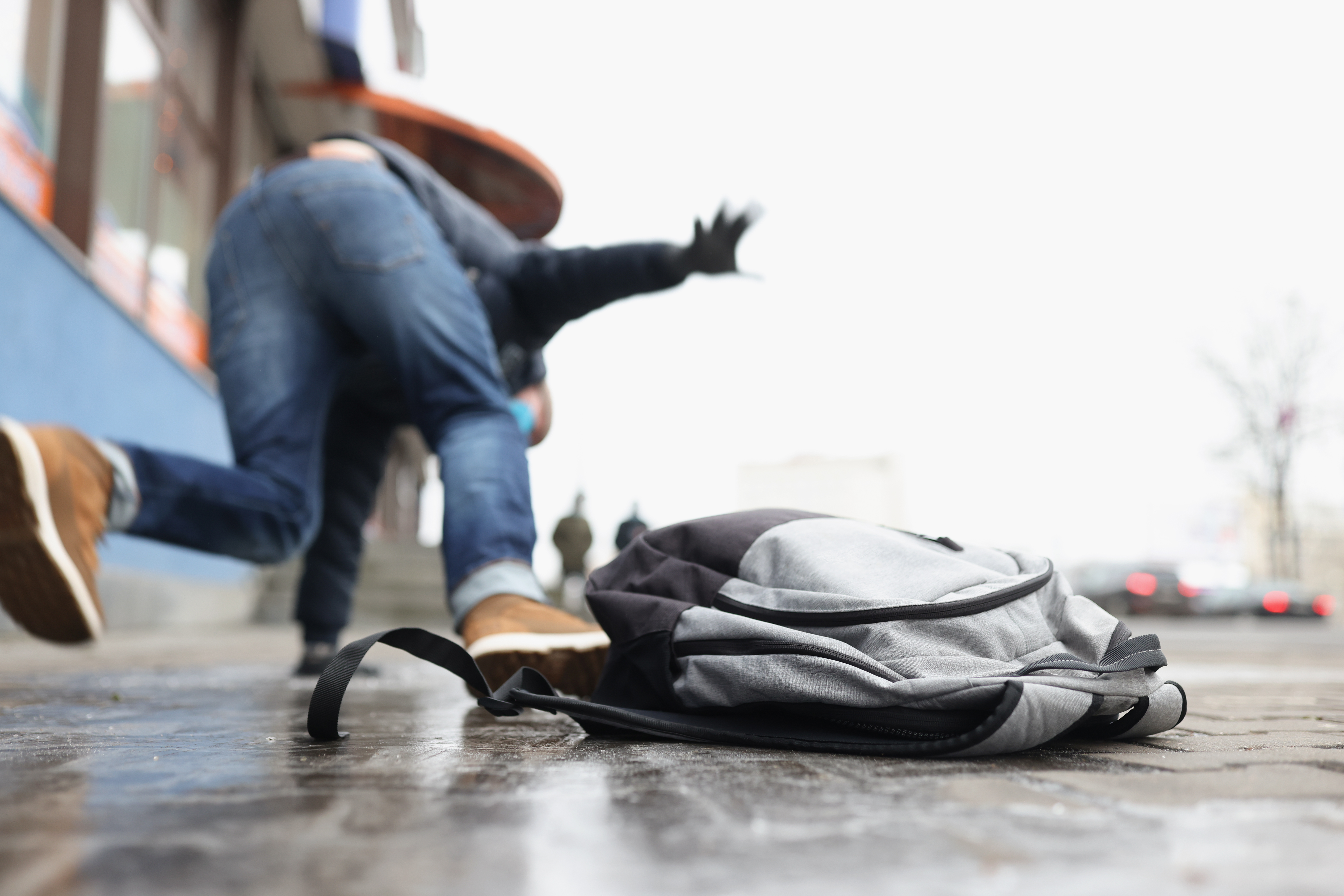 A person drops their backpack as they fall to the ground after slipping on an icy sidewalk outside of a business. 