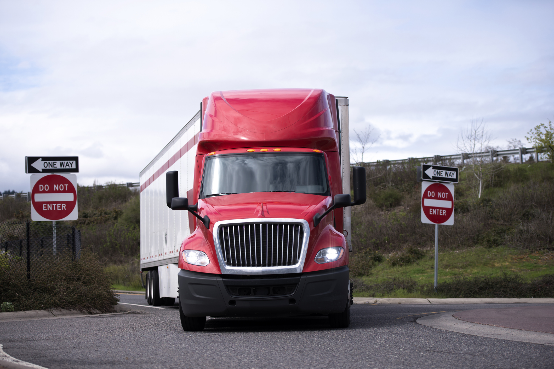 A semi-truck with a bright red cab takes a wide right turn onto a street after coming off of an interstate exit.